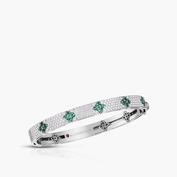 A white gold bangle bracelet featuring pavé-set round stones and green emerald-cut diamond floral motifs, with a small red gemstone accent on the inner band.